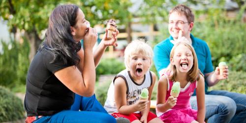 Eine Familie, Mutter, Vater und zwei Kinder, sitzen auf einer Bank. Jede Person isst ein Eis.