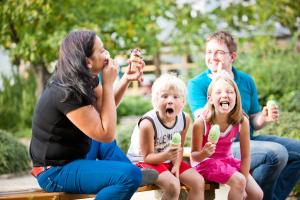 Eine Familie, Mutter, Vater und zwei Kinder, sitzen auf einer Bank. Jede Person isst ein Eis.